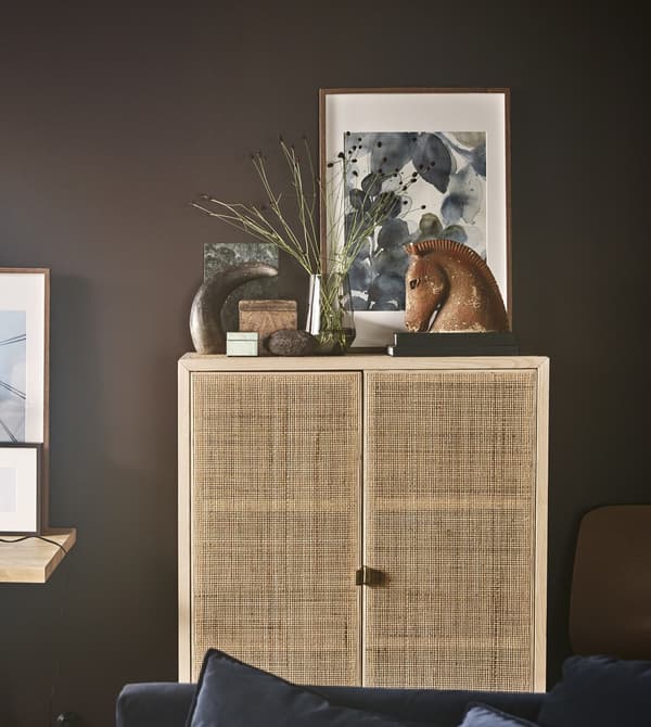 Un armario IKEA STOCKHOLM de madera maciza y ratán sostiene artículos decorativos, como un florero con flores y una escultura de madera.