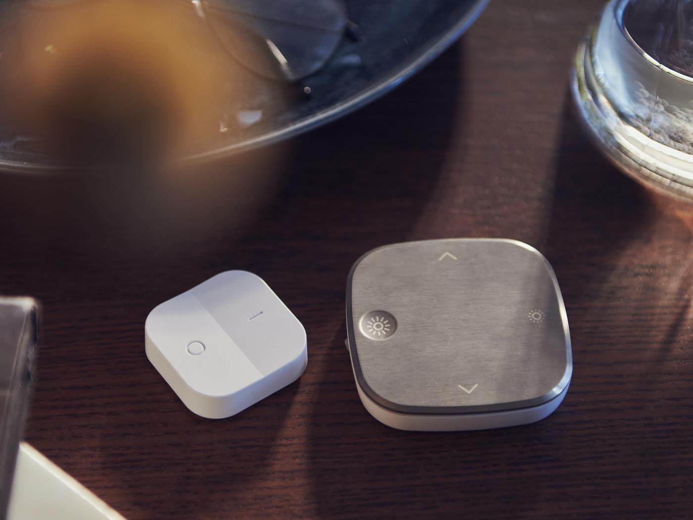 Un atenuador inalámbrico TRÅDFRI blanco y un mando a distancia STYRBAR, uno junto al otro, en una mesa marrón con un vaso de agua.