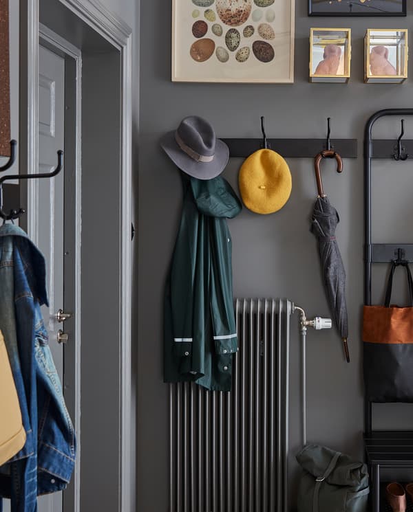 Un attaccapanni nero a tre ganci, montato a parete sopra un calorifero, con appesi un ombrello, due cappelli e un impermeabile - IKEA