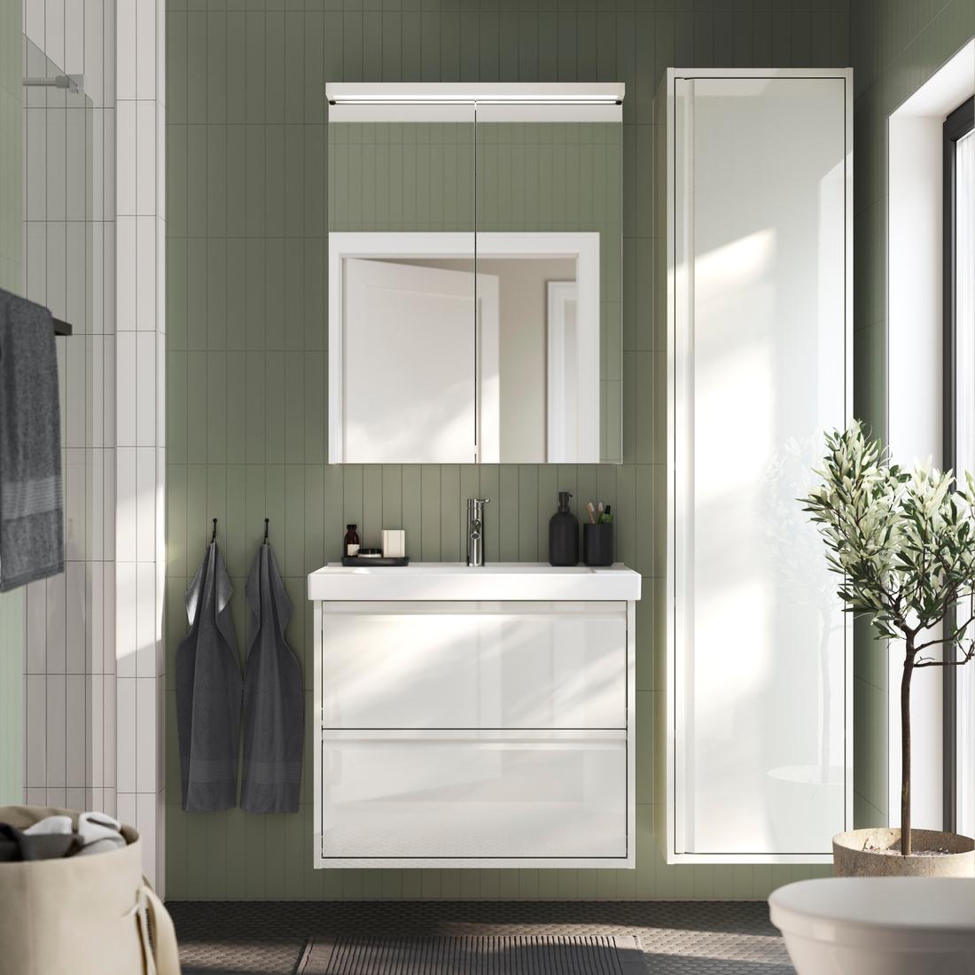 Un bagno ÄNGSJÖN lucido bianco visto frontalmente completo di lavabo in ceramica bianco, mobile per lavabo con due cassetti, pensile a specchio con due ante e un mobile a colonna sospeso con anta. Di fianco un box doccia e un cestino per la biancheria.