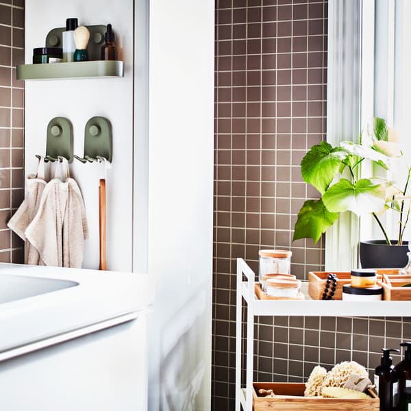 Un bagno con le piastrelle grigie con un lavabo bianco, un mobile alto bianco, un carrello bianco contenente vassoi e barattoli e una pianta in vaso.