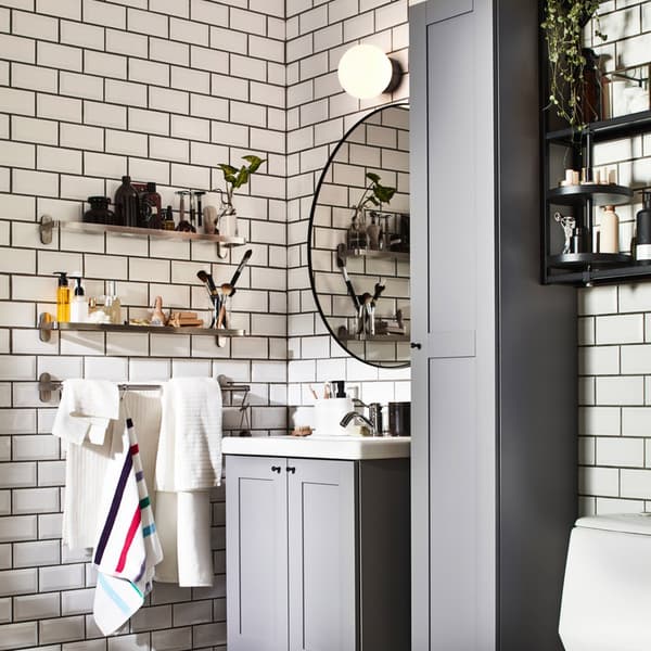 Un bagno con piastrelle bianche e fughe nere. Ci sono mobili grigi, uno specchio tondo e una luce accesa a parete.