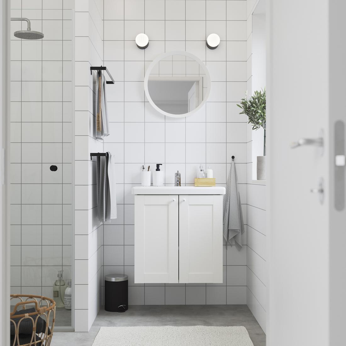 Un bagno EHNET bianco visto frontalmente completo di lavabo in ceramica bianco, mobile per lavabo con due ante, specchio bianco rotondo. Di fianco un box doccia e un cestino per la biancheria.