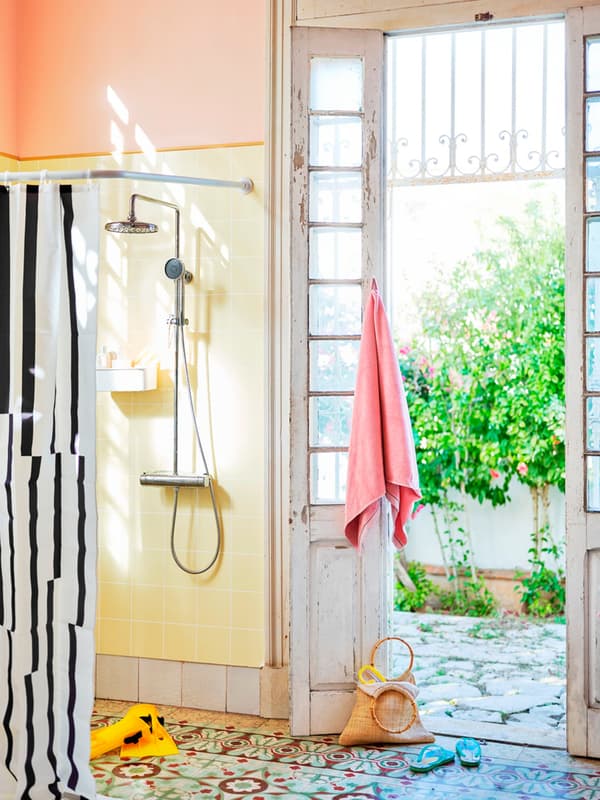 Un bagno molto illuminato con piastrelle colorate. A sinistra, c'è una doccia con tendina, alla porta finestra è appeso un asciugamano rosa e sul pavimento ci sono una borsa e delle infradito.