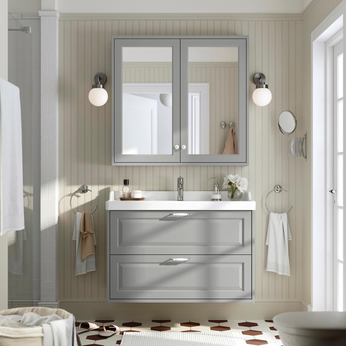 Un bagno TÄNNFORSEN grigio visto frontalmente completo di lavabo in ceramica bianco, mobile per lavabo con due cassetti, pensile a specchio con due ante. Illuminazione a parete sferica ai lati del pensile.