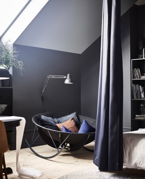 Un balancí IKEA PS 2017 crea un racó de lectura en un dormitori amb parets negres.