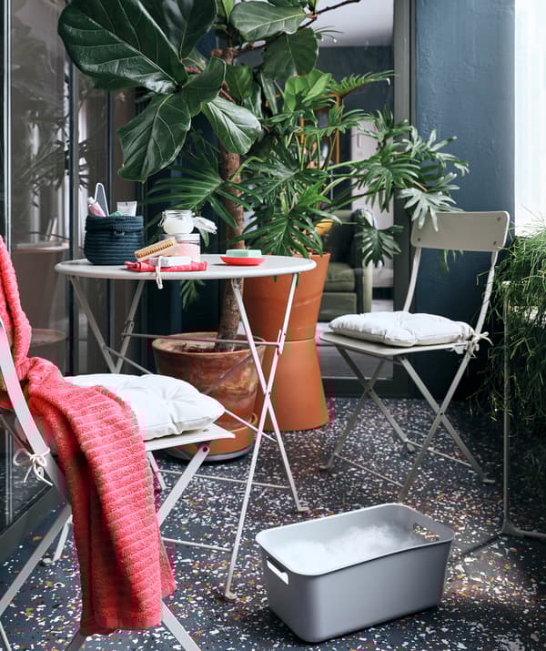 Un balcon avec de grandes plantes en pot, un petit coin où s’asseoir et un bain de pieds dans une boîte GRUNDVATTNET à côté d’une chaise SALTHOLMEN.