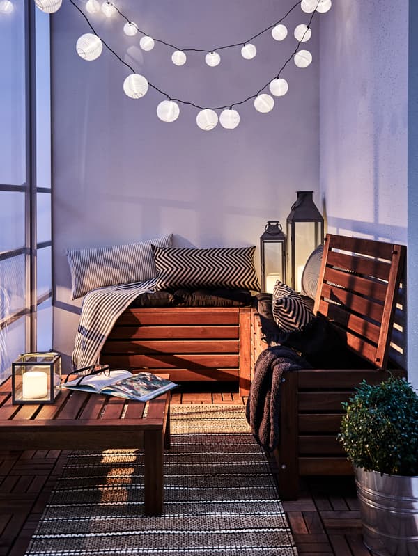 Un balcon avec du mobilier d’extérieur en bois complété par des coussins, des lampes, des luminaires en suspension et une plante.