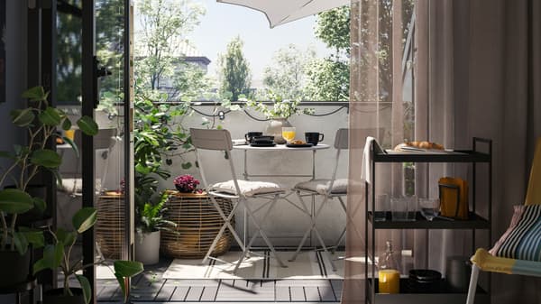 Un balcon avec une table d’extérieur SUNDSÖ grise et deux chaises d’extérieur grises sur un tapis, plus une table basse, des plantes et un parasol.