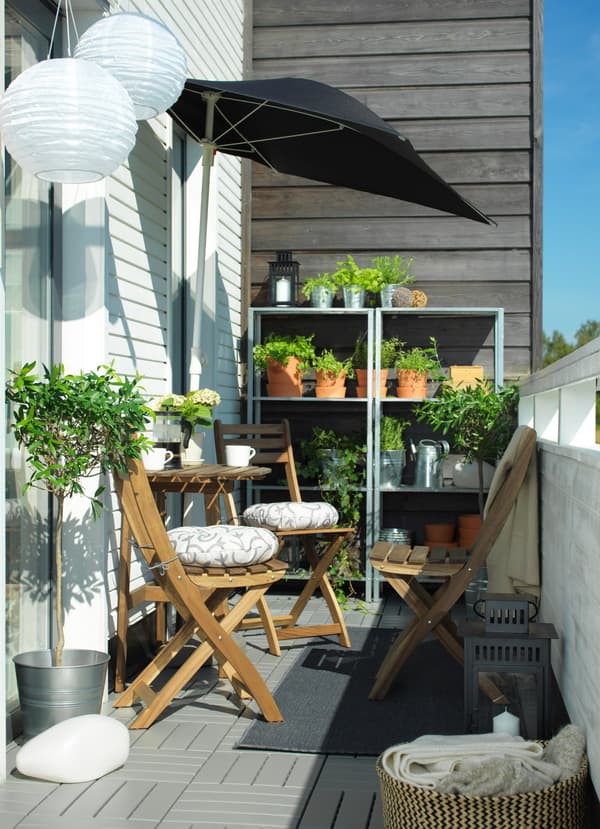 Un balcon étroit avec une table et des chaises en bois au soleil. Des étagères garnies de rangées de plantes sont visibles en arrière-plan.