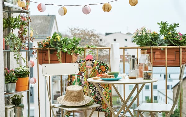 Un balcon urbain décoré de jardinières et d’une guirlande lumineuse, meublé d’une étagère de plantes et de deux chaises et une table style bistrot, sur laquelle est servi un en-cas.