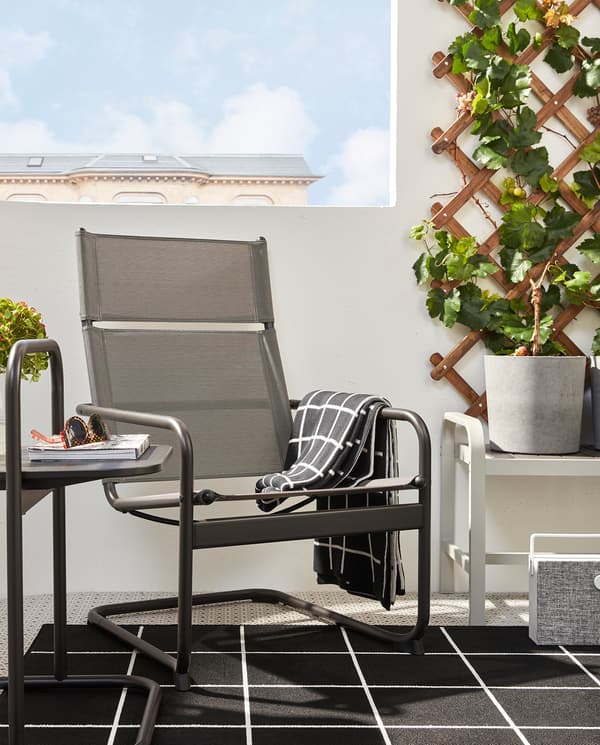 Un balcone con poltrona e tavolino HUSARÖ grigio scuro su un tappeto nero e bianco con accanto una panca con un portavaso - IKEA