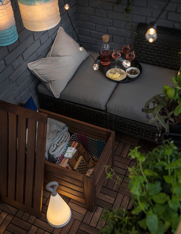 Un balcone con un contenitore ÄPPLARÖ aperto che svela dei giochi e dei plaid. Nell'angolo si trova una seduta modulare KUNGSHOLMEN con dei cuscini e materassi e sopra di questi è posato un vassoio con bevande e snack. Sulla destra si intravedono delle piante e dal soffito pendono delle lampade. 