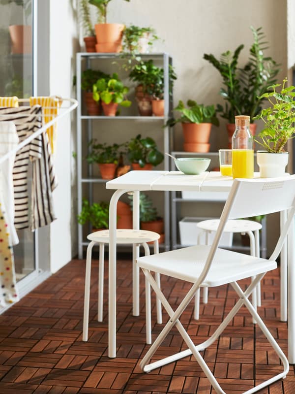 Un balcone con un tavolino e sedie da esterno bianco. Si intravede uno stendibiancheria aperto e sullo sfondo ci sono scaffali con piante in vaso.
