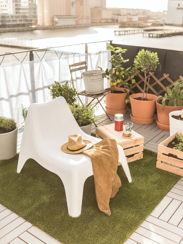 Un balcone con una poltrona bianca SKARPÖ su cui sono appoggiati un indumento e un cappello di paglia. Attorno alcuni vasi con piante e contenitori in legno, di cui uno usato a mo' di tavolino.
