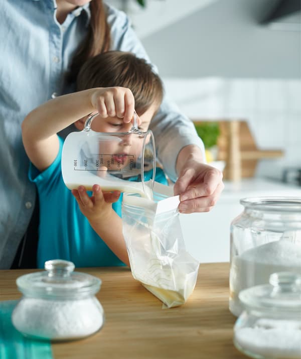 Un bambino che con l'aiuto della mamma sta preparando il gelato versando un liquido in un sacchetto di plastica.