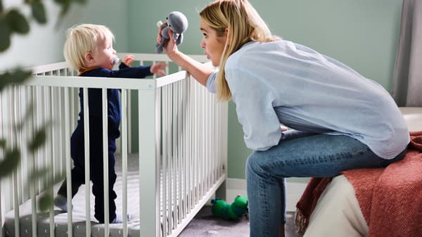Un bambino con un ciuccio e vestiti blu scuro è in piedi in un lettino GULLIVER bianco e un adulto si china verso di lui con un peluche grigio - IKEA.