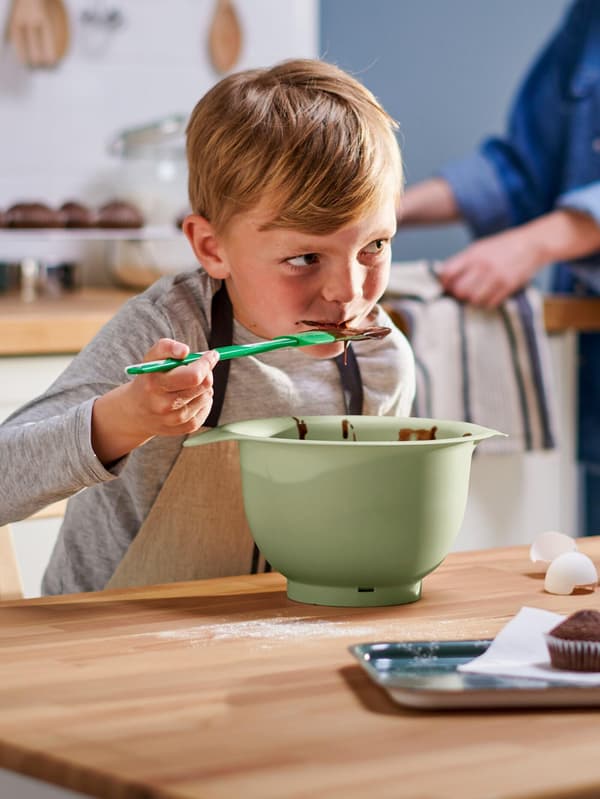 Un bambino con un grembiule MARIATHERES mangia l'impasto di una torta al cioccolato da una ciotola verde servendosi di una spatola GUBBRÖRA verde/bianco.