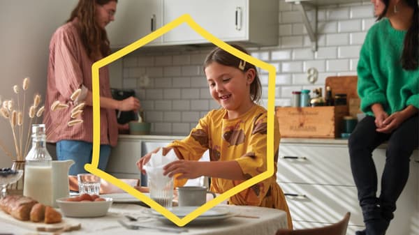 Un bambino con un vestito giallo mette un tovagliolo in un bicchiere su un tavolo, mentre un’altra persona in una cucina luminosa mescola gli ingredienti in una ciotola con piastrelle bianche.