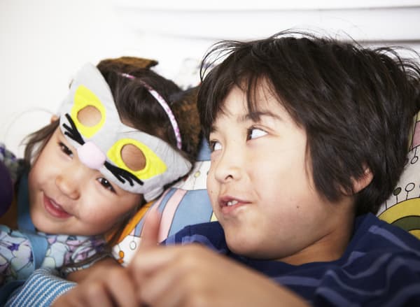 Un bambino e una bambina con una maschera da gatto sollevata sulla fronte, seduti sul divano a parlare.