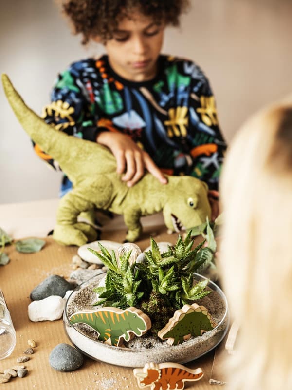 Un bambino gioca con un peluche di un dinosauro su un tavolo con una pianta al centro e diverse sagome di altri dinosauri. 