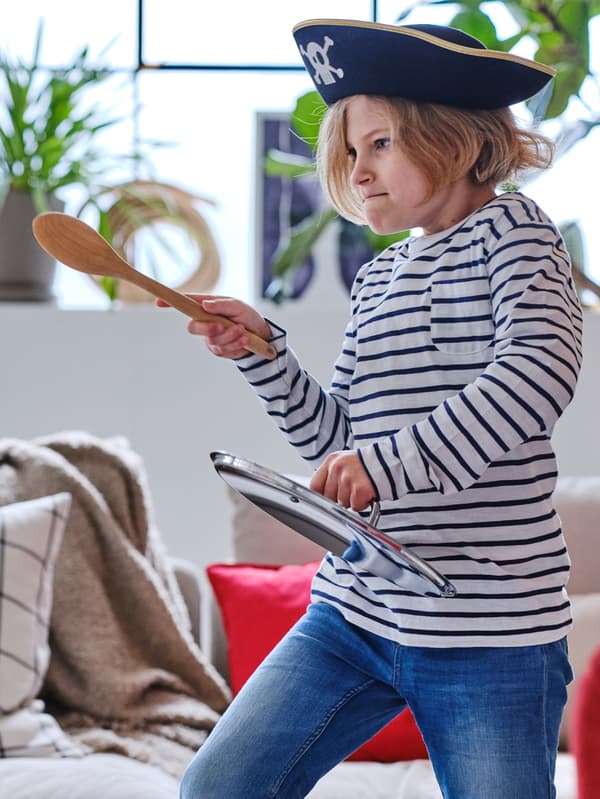 Un bambino in jeans, maglia a righe e cappello da pirata gioca con un cucchiaio in legno RÖRT e un coperchio per casseruola simulando un duello alla spada.