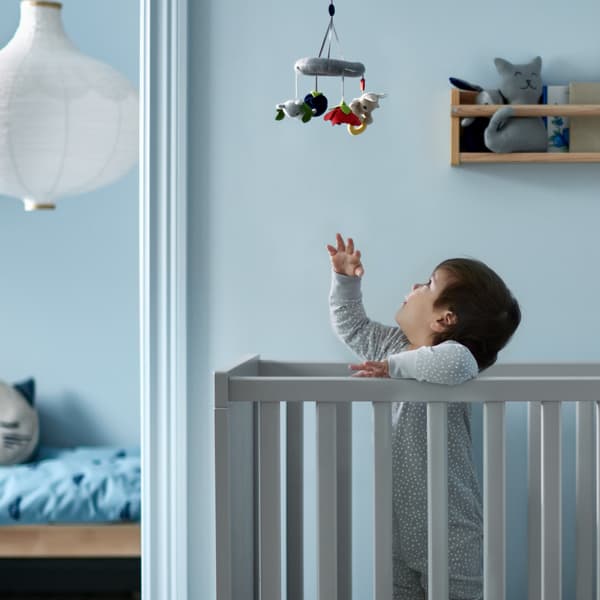 Un bambino in piedi in un lettino SUNDVIK grigio allunga una mano verso la giostrina GULLIGAST multicolore appesa in alto - IKEA.