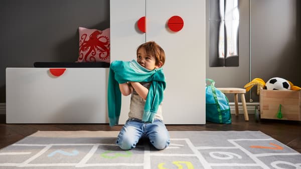 Un bambino si veste in ginocchio su un tappeto grigio, di fronte a una panca bianca con contenitore per giocattoli e a un guardaroba bianco con un elemento estraibile.