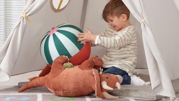 Un bambino sta giocando all'interno di una tenda HÖVLIG con un pallone e un peluche a forma di dinosauro.