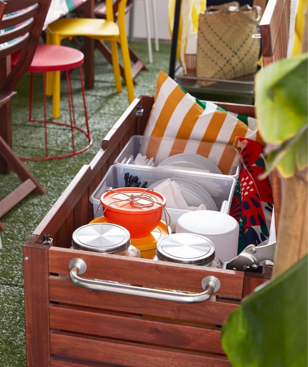 Un banc de rangement IKEA ÄPPLARÖ en bois d’acacia teinté brun à utiliser en extérieur, rempli d’objets pour une fête.