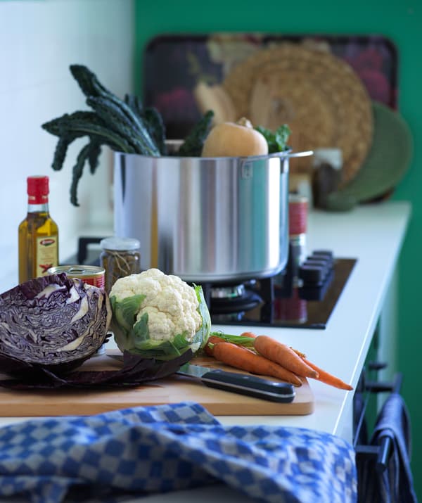 Un bancone di una cucina su cui poggiano diverse verdure. Sul fuoco c'è una pentola in acciaio con all'interno diversi alimenti. Sullo sfondo si intravedono vassoi e sottopiatti.