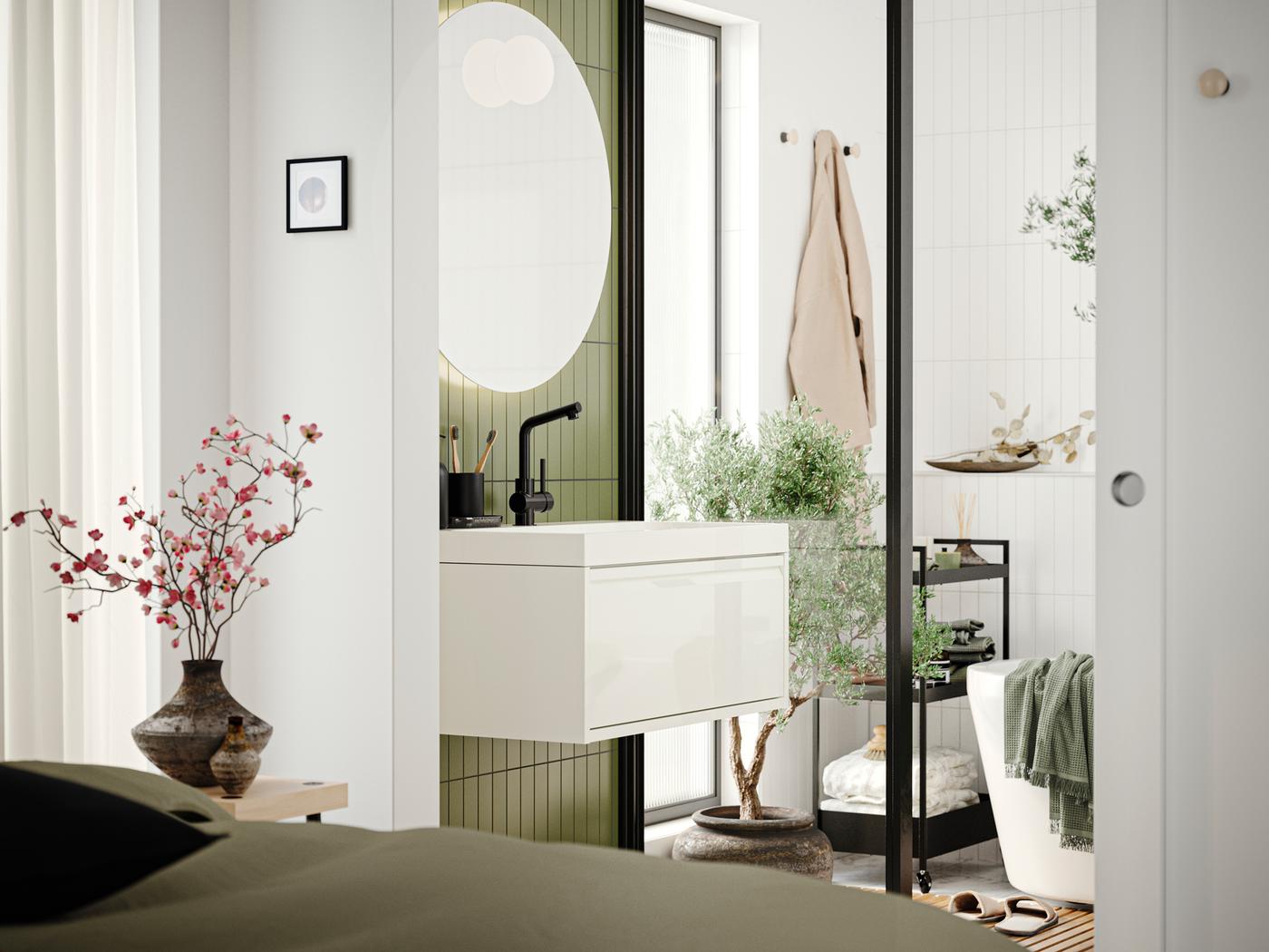Un baño con un mueble de lavabo con cajón ÄNGSJÖN en blanco de alto brillo, un espejo y una bañera visto desde un dormitorio.