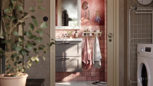 Un baño de azulejos rosas con un gabinete ENHET gris con cajones, un espejo blanco y dos toallas colgadas de una barra con ganchos.