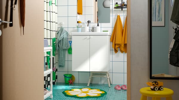 Un baño familiar con mobiliario ENHET y un suelo verde con una alfombra KÄRRKNIPPROT, y ganchos de pared a diferentes alturas.