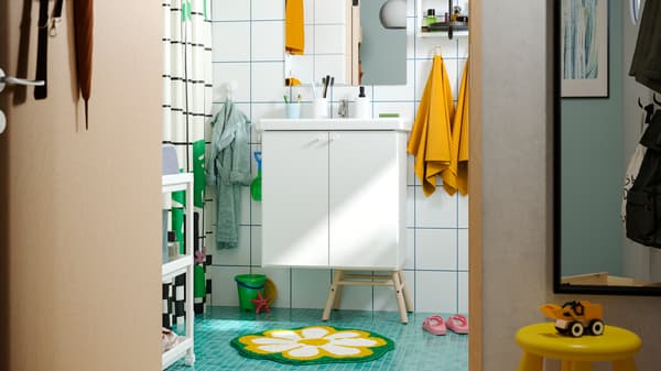Un baño familiar con piso verde, toallas amarillas, tapete floral, ganchos de pared en diferentes alturas y muebles ENHET blancos.