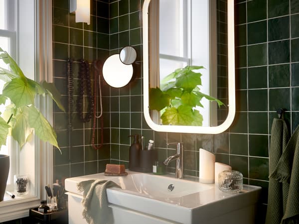 Un bany amb un mirall blanc amb il·luminació integrada sobre un moble de lavabo, tovalloles per a convidats de color verd clar i una planta.