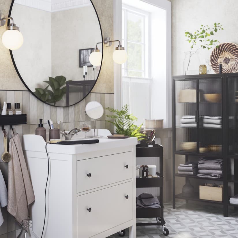 Un bany on hi ha un mirall rodó, dos llums de paret i un moble de lavabo de color blanc amb dos calaixos i poms negres de la sèrie HEMNES.