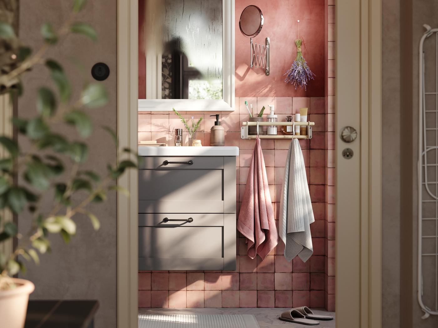 Un bany rosa amb un armari ENHET gris amb un lavabo blanc i un mirall a sobre. Hi ha tovalloles penjades als ganxos del costat.