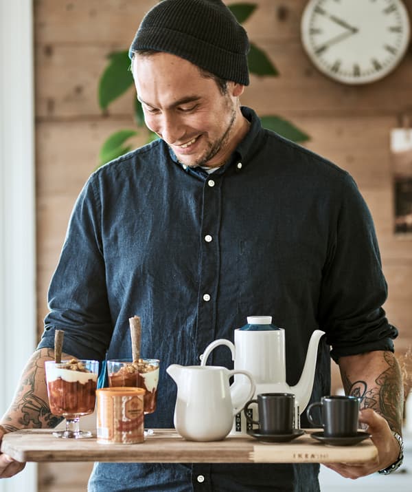 Un bărbat care poartă o căciulă pe cap folosește un tocător pe post de tavă pentru a duce un ceainic cu cafea, cești de espresso și două pahare cu granola.