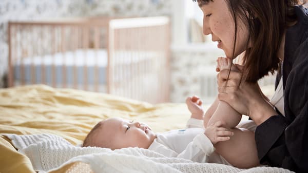 Un bebé feliz sobre una manta GULSPARV blanca encima de una cama junto a su madre, que le sujeta los pies y le sonríe.