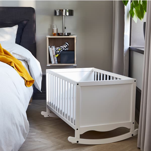 Un berceau blanc SOLGUL à côté d’une fenêtre de chambre à coucher et un lit-sommier gris foncé avec du linge de lit rayé et gris et une couverture jaune foncé.
