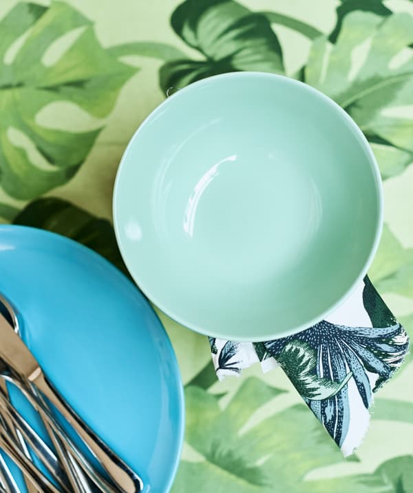 Un bol vert clair à côté d’une assiette turquoise sur laquelle sont posés des couverts, le tout sur une table recouverte d’un tissu à motif de feuilles vertes.