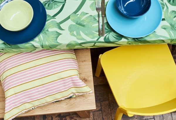 Un bord de table couvert d’un tissu à imprimé de feuilles vertes, une chaise jaune et un banc en chêne avec un coussin jaune à rayures.