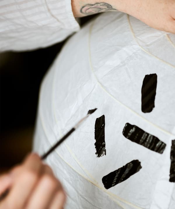 Un bras au poignet tatoué tient un abat-jour en papier blanc pendant qu'on y peint des traits noirs.