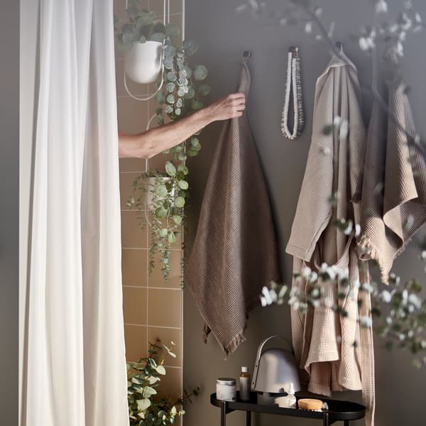 Un bras masculin, derrière un rideau de douche BASTSJÖN blanc, se tend vers une serviette brune dans une salle de bain confortable avec quelques plantes.