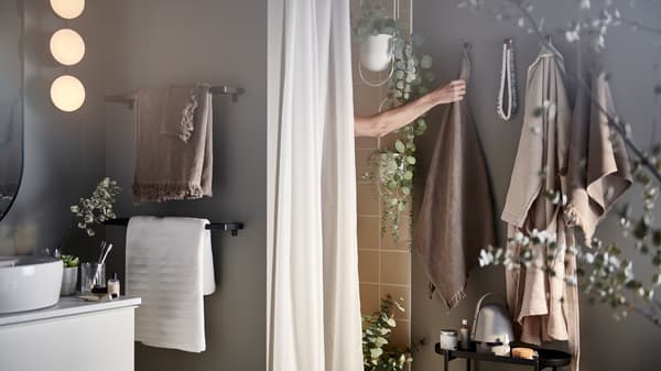 Un bras sortant de la douche pour saisir une serviette brun/gris clair sur un crochet, plus un rideau de douche et des plantes artificielles en pot.