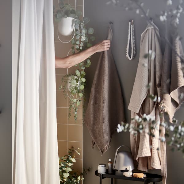 Un bras sortant de la douche pour saisir une serviette brun/gris clair sur un crochet, plus un rideau de douche et des plantes artificielles en pot.