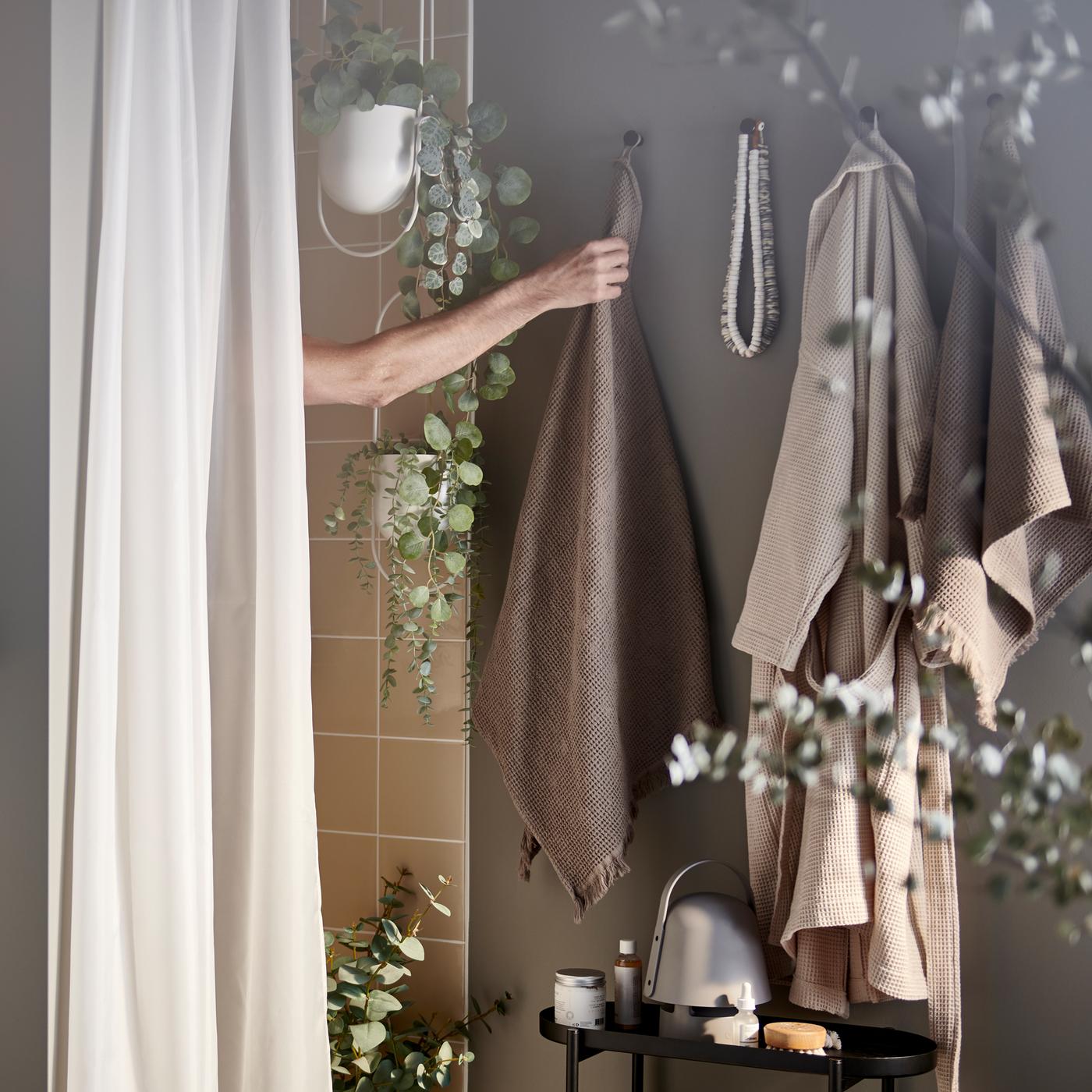 Un bras tendu d’homme attrape une serviette brune derrière un rideau de douche BASTSJÖN blanc dans une salle de bains spa chaleureuse et ornée de quelques plantes.