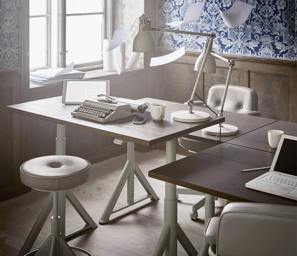 Un bureau à hauteur réglable et une chaise placés à proximité d’une fenêtre, dans un espace décoré de papier peint à motif bleu.