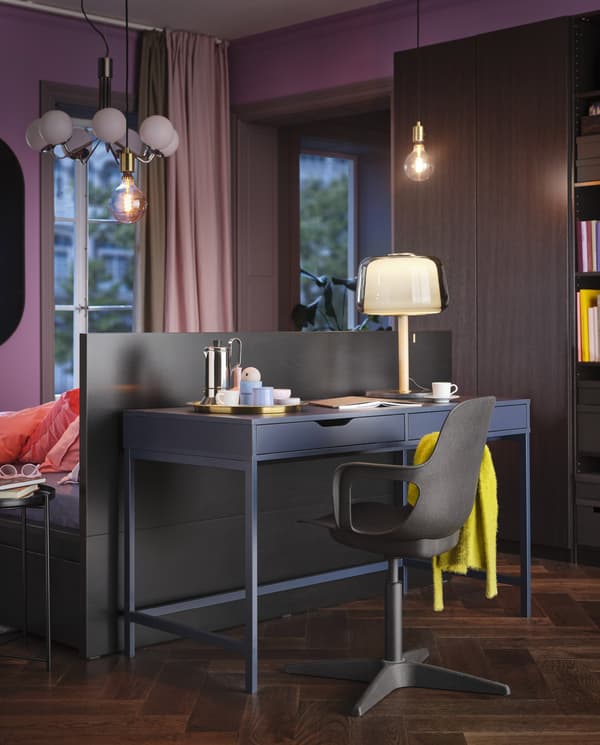 Un bureau ALEX bleu, une lampe de table allumée et une chaise pivotante noire avec un plaid jaune, à l’arrière d’une tête de lit.